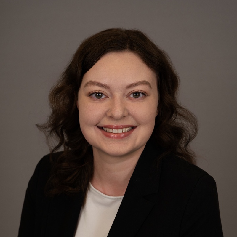 Professional headshot of a smiling woman with shoulder-length wavy brown hair, wearing a black blazer over a white top against a neutral gray background.