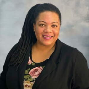 Professional Headshot Of A Smiling Black Woman With Long Locs, Wearing A Black Blazer Over A Floral Top Against A Soft Gray-blue Background