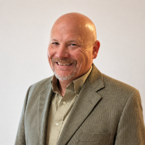 Professional Headshot Of A Smiling Bald Man With A Salt-and-pepper Goatee, Wearing A Tan Patterned Blazer And Light Olive Shirt Against A Plain Light Background.
